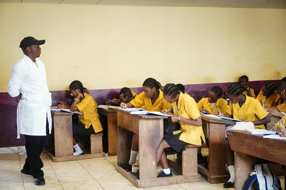 Teacher in a white coat walking between rows of students writing in class